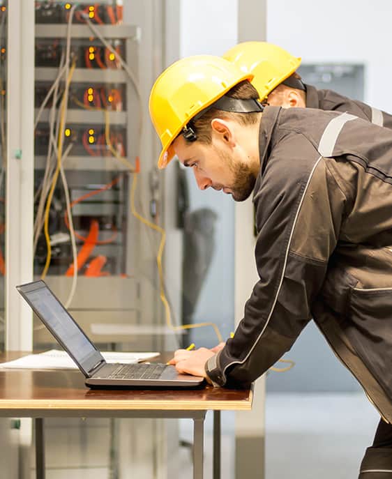 Two men in hard hats collaborate on a laptop at a construction site.