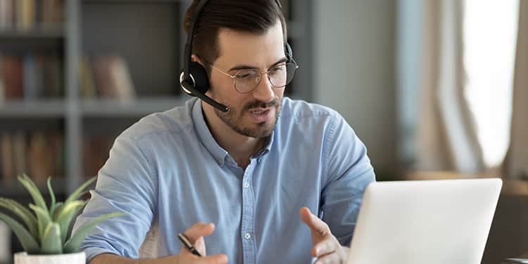 Man with glasses and headset sits at a desk, speaking during a video call on a laptop.