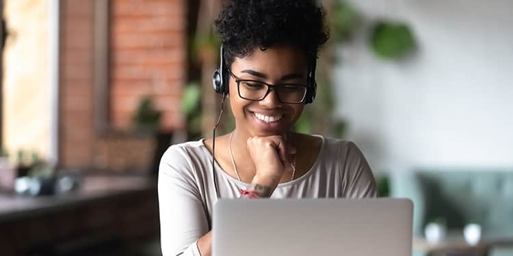 A woman wearing glasses and a headset smiles while working on her laptop.