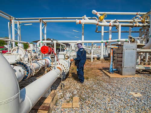 A man in a blue suit stands beside industrial pipes.