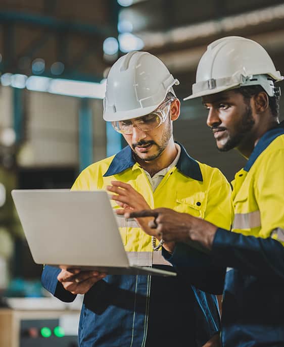 Two workers wearing hard hats and safety vests examining a laptop together.