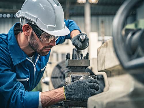 A man wearing a hard hat and safety glasses operates machinery in a workshop setting.