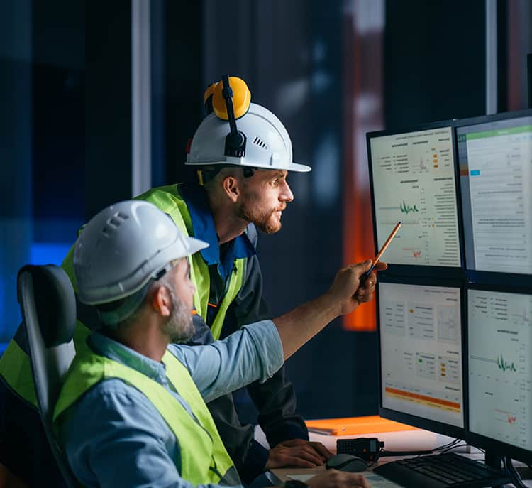 Two men wearing hard hats focused on computer screens, working together.